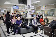 Кілька людей сидять у черзі на стільцях у великому залі з яскравим освітленням, ймовірно, чекають на прийом у ЦНАП