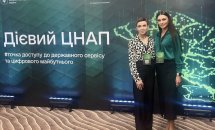 Дві жінки фотографуються перед зеленою стіною з написами конференції.