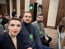 Селфі трьох людей у конференц-залі, двоє чоловіків і жінка на передньому плані.