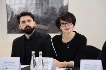 Чоловік із бородою та жінка в окулярах сидять за столом із табличками, перед ними пляшки з водою.