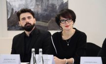 Чоловік із бородою та жінка в окулярах сидять за столом із табличками, перед ними пляшки з водою.