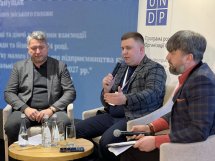 Троє чоловіків сидять. Один проголошує доповідь, а інші його слухають