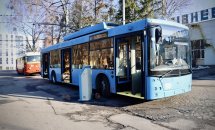 На дворі стоїть синій тролейбус. Збоку стоїть термінал, на задньому фоні стоїть червоний тролейбус.