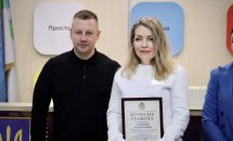 Чоловік та жінка стоять у приміщені  ЦНАПу в місті Рівному. Жінка тримає у руках грамоту.