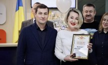 Чоловік та жінка стоять у приміщені. Жінка тримає у руках почесну грамоту.