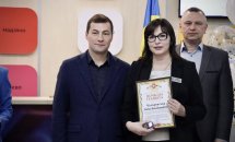 Два чоловіки та жінка стоять у приміщені. Жінка тримає у руках почесну грамоту.