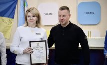 Чоловік та жінка стоять у приміщені. Жінка тримає у руках почесну грамоту.