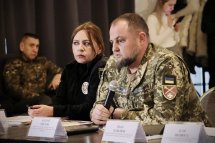 Чоловік у військовій формі тримає мікрофон, біля нього сидить жінка. На задньому фоні люди.