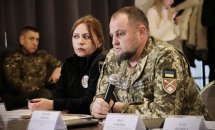 Чоловік у військовій формі тримає мікрофон, біля нього сидить жінка. На задньому фоні люди.