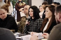 Жінка сидить за столом та тримає у руках мікрофон. На задньому фоні інші учасники зустрічі.