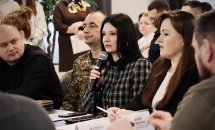 Жінка сидить за столом та тримає у руках мікрофон. На задньому фоні інші учасники зустрічі.