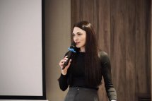 Жінка з довгим волоссям тримає в руках мікрофон та проголошує доповідь.