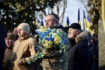 Чоловік у військовій формі триває букет з жовтих та блакитних хризантем. На задньому фоні стоять люди на дворі.