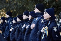 Працівники поліції тримаючи руку на серці, стоять на дворі. Вони у формі чорного кольору та у головних уборах.