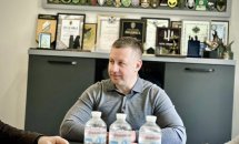 Чоловік у сірому светрі сидить за столом. За його спиною шафа нагородами, грамотами та емблемами.