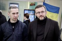 Два чоловіки стоять у приміщенні. На задньому фоні прапор України та жінка поруч з інформаційним терміналом