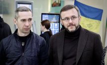 Два чоловіки стоять у приміщенні. На задньому фоні прапор України та інформаційний термінал.