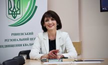 Жінка з коротким темним волоссям сидить за столом у світлому піджаку.