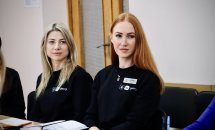 Дві жінки сидять за столом. Вони одягненні у чорні брендовані кофти. На столі лежать записники.