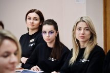 Дві жінки та дівчина в окулярах сидить за столом у чорних кофтах, уважно слухаючи лектора.