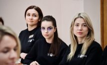 Дві жінки та дівчина в окулярах сидить за столом у чорних кофтах, уважно слухаючи лектора.