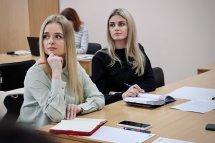Дві жінки з світлим волоссям сидять за столом. Одна жінка одягнена у чорну кофту, а інша - у блузу зеленого кольору. Вони уважно слухають лектора. На столі лежать записники та роздаткові матеріали.