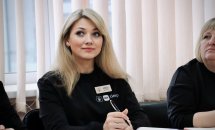 Жінка з білим волосся у чорній кофті з бейджем сидить за столом. У руках вона тримає кулькову ручку.
