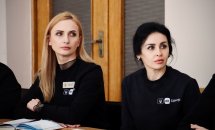 Дві жінки у чорних кофтах з бейджами сидять за столом, уважно слухаючи доповідача.