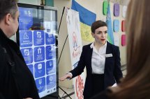 Жінка у діловому одязі стоїть біля інформаційного терміналу та щось розповідає.
