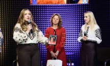 Жінка з довгим волоссям тримає подяку у руках та щось говорить, біля неї стоять дві жінки.