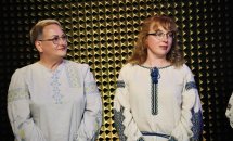Дві жінки в окулярах та вишитих сорочках стоять на чорному фоні.