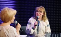 Жінка в окулярах та вишиванці тримає мікрофон, біля неї сидить жінка.