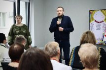 Чоловік з мікрофоном стоїть перед групою людей у приміщенні.