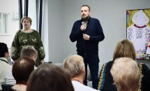 Чоловік з мікрофоном стоїть перед групою людей у приміщенні.