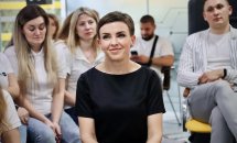 Жінка в чорному одязі сидить на заході серед інших учасників та усміхається.