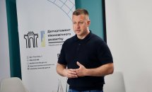 Чоловік в темному одязі стоїть у приміщенні, на фоні - банер та два стільці