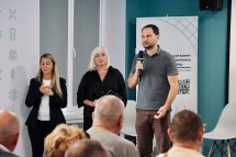 Чоловік говорить у мікрофон перед аудиторією, біля нього стоять дві жінки.