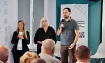 Чоловік говорить у мікрофон перед аудиторією, біля нього стоять дві жінки.