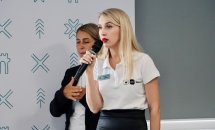 Жінка зі світлим волоссям тримає мікрофон, позаду неї стоїть жінка.