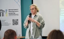 Жінка зі світлим волоссям, в окулярах, тримає мікрофон у руках.