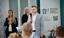 Чоловік у діловому одязі говорить у мікрофон перед аудиторією, позаду — дві жінки.
