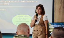 Жінка з темним волоссям говорить у мікрофон перед аудиторією.