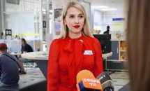 Жінка зі світлим волоссям, у червоній сукні, дає інтерв'ю у приміщенні.