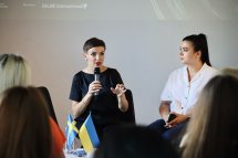 Жінка з коротким волоссям говорить у мікрофон, біля неї сидить жінка.