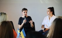 Жінка з коротким волоссям говорить у мікрофон, біля неї сидить жінка.