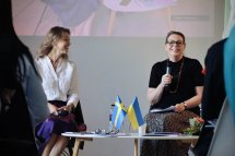 Дві жінки сидять на сцені під час заходу, одна з мікрофоном. На столику перед ними прапорці.