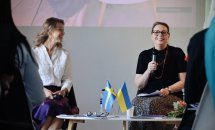 Дві жінки сидять на сцені під час заходу, одна з мікрофоном. На столику перед ними прапорці.