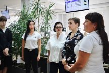 Група людей стоїть у приміщенні, поруч табло з номерами черги та зелені рослини.