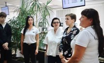 Група людей стоїть у приміщенні, поруч табло з номерами черги та зелені рослини.