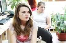 Жінка з довгим волоссям сидить у приміщенні, позаду неї сидить ще одна жінка.